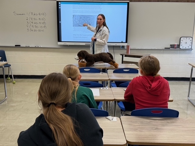 A speaker is discussing her career with a group of students sitting in their desks. There is a presentation on the screen.