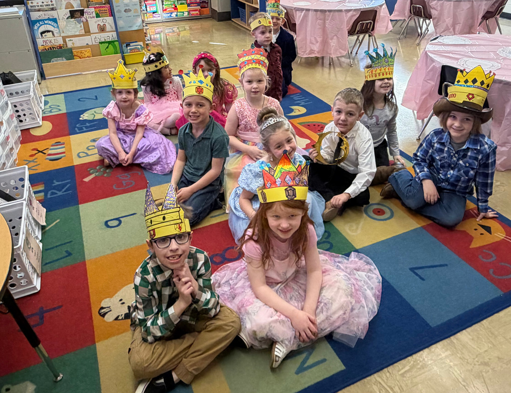 The students are sitting on the carpet. They are wearing fancy clothing and a crown.