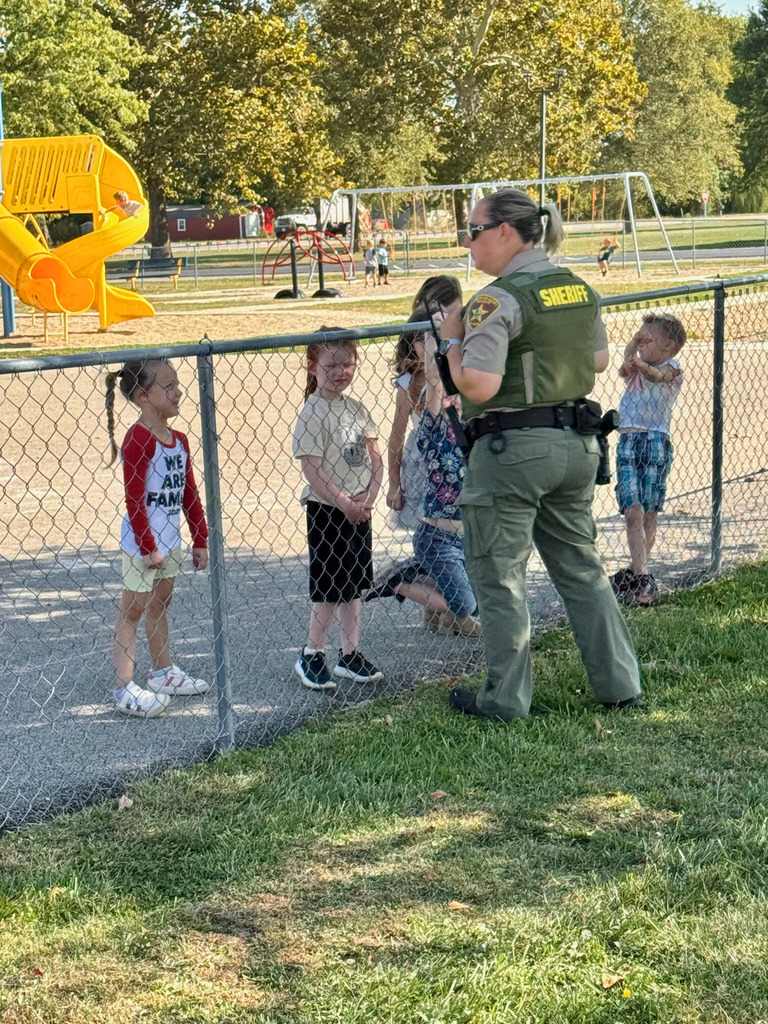 Officer E is standing at the playground fence. She is talking to four students at recess. 