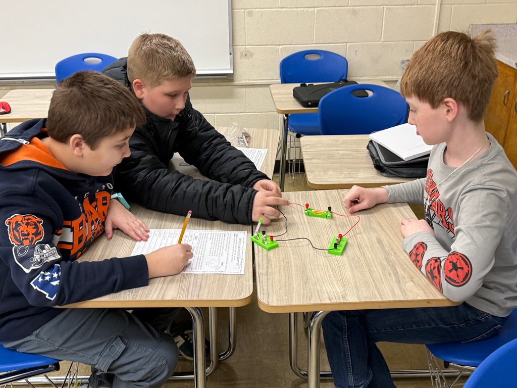 Students are in their desks with their circuit. They are adding an everyday object to the circuit to see if the bulb lights,  They are writing their results on their lab sheet. 