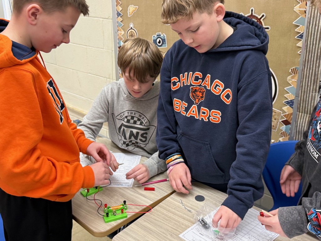 Students are in their desks with their circuit. They are adding an everyday object to the circuit to see if the bulb lights,  They are writing their results on their lab sheet. 