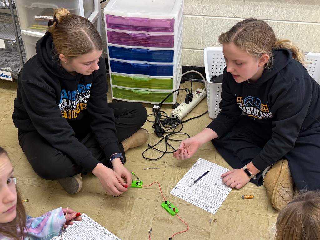 Students are on the floor with their circuit. They are adding an everyday object to the circuit to see if the bulb lights,  They are writing their results on their lab sheet. 