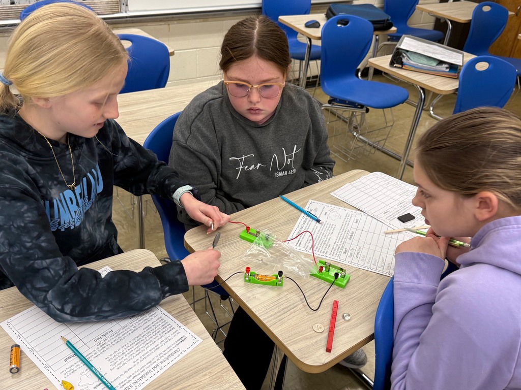 Students are in their desks with their circuit. They are adding an everyday object to the circuit to see if the bulb lights,  They are writing their results on their lab sheet. 