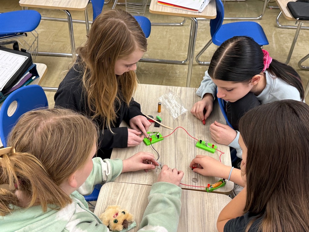 Students are in their desks with their circuit. They are adding an everyday object to the circuit to see if the bulb lights,  They are writing their results on their lab sheet. 