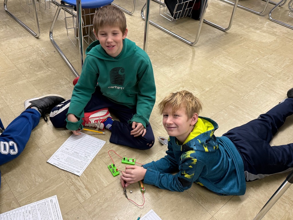 Students are on the floor with their circuit. They are adding an everyday object to the circuit to see if the bulb lights,  They are writing their results on their lab sheet. 