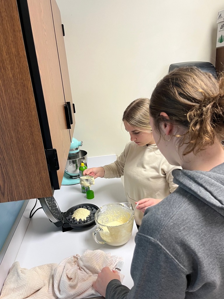 A student is dipping batter and pouring it onto the  waffle iron. Another student is looking on. 