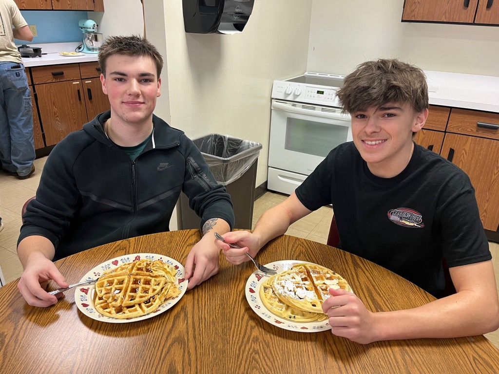 Two students are sitting at the table with plates of waffles with toppings like powdered sugar and syrup. 