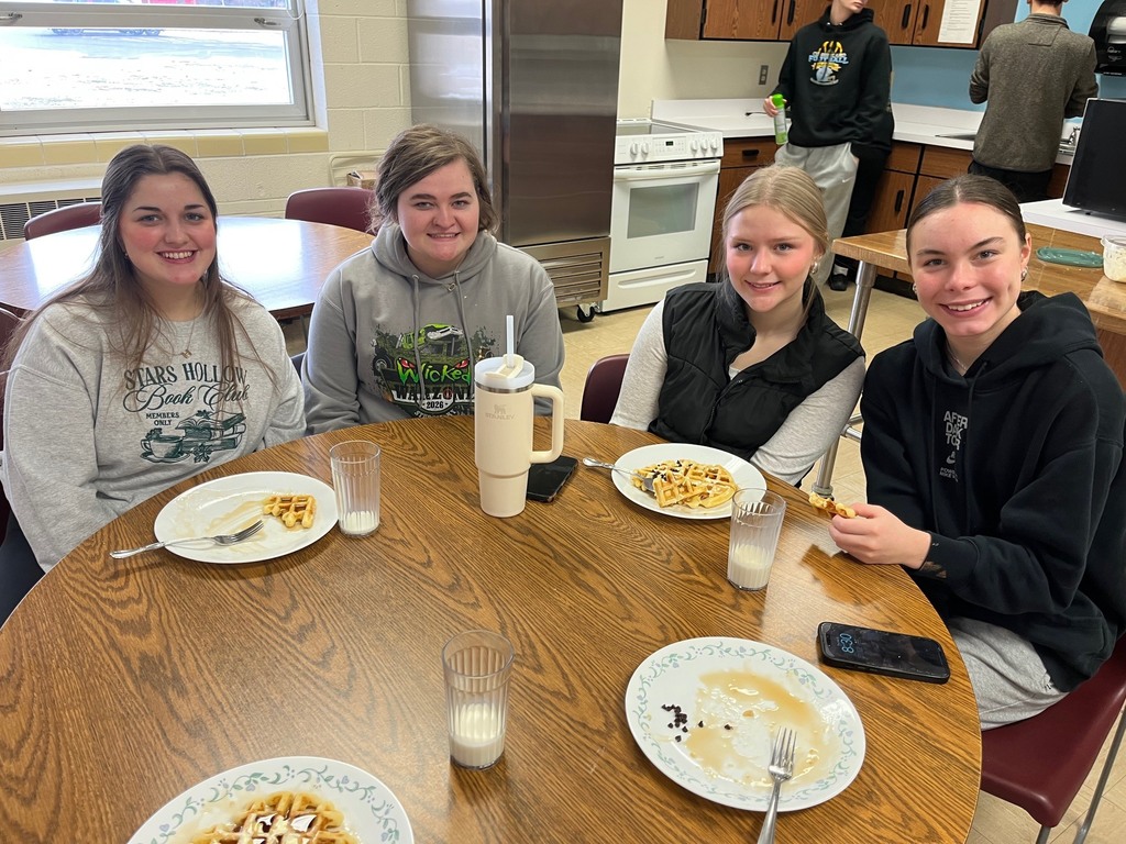 Four students are sitting at the table with plates of waffles with toppings like powdered sugar and syrup. 