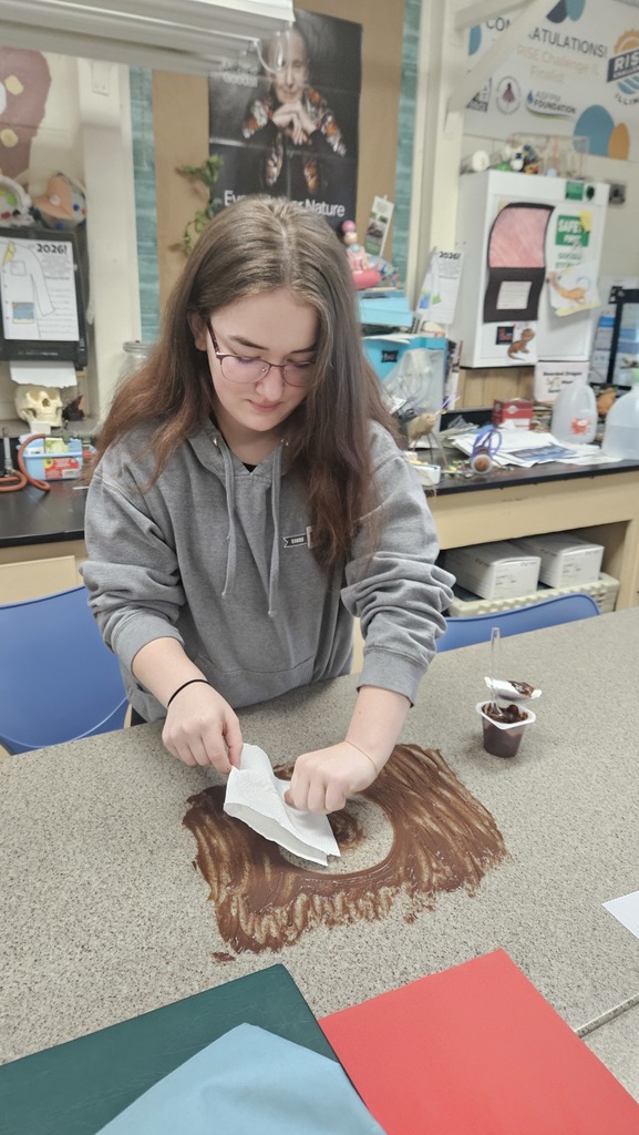 With pudding smeared on the table, a student is using circular motions to wipe up the pudding. 