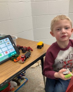 A student is using an ipad for words. The student is playing with toy cars and trucks.