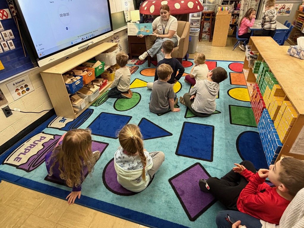 Eight students are sitting on a shape on the carpet. The students are listening to their teacher read a story about getting a pet. 