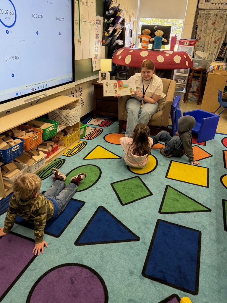 Three students are sitting on a shape on the carpet. The students are listening to their teacher read a story about getting a pet. 