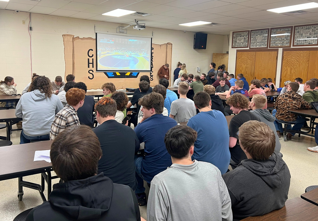 Pictured are a group of about 30 students watching the wrestling match online in the cafeteria. The match is being projected on the  white screen. 