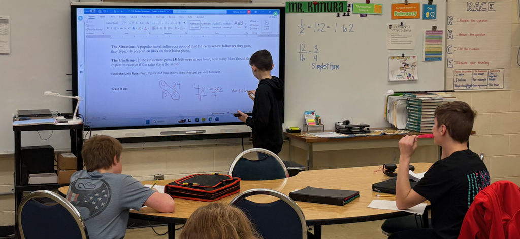 A student is at the white board working out a problem of ratios and proportions. There are three students at their desks working the problem with him. 
