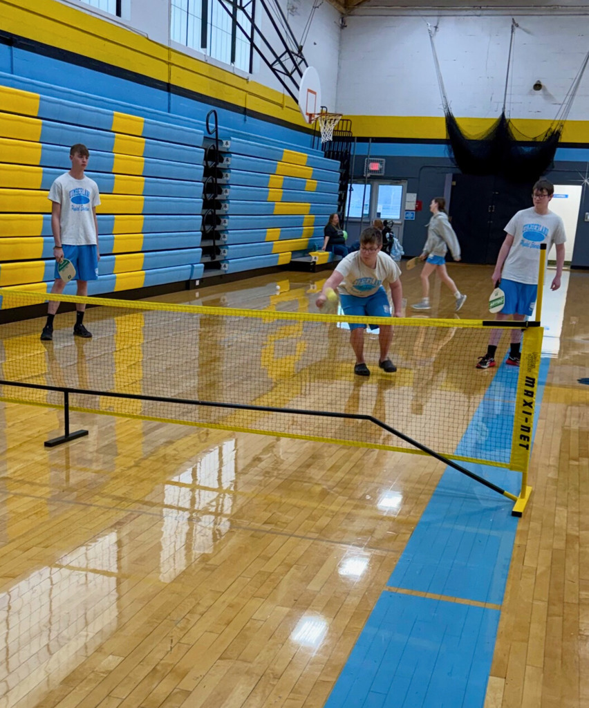 Three students are standing behind the net. One student is underhand hitting the ball back over the net. 