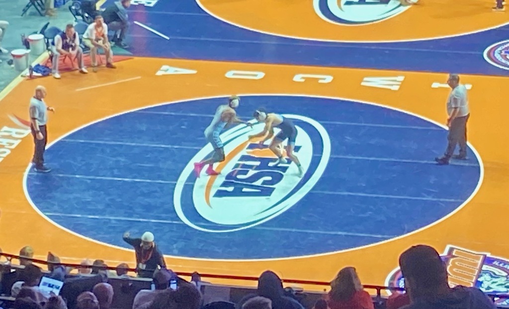 OM and another wrestler are on the center of the mat. They are facing each other with arms outstretched. There are two officials on either side of the mat. 