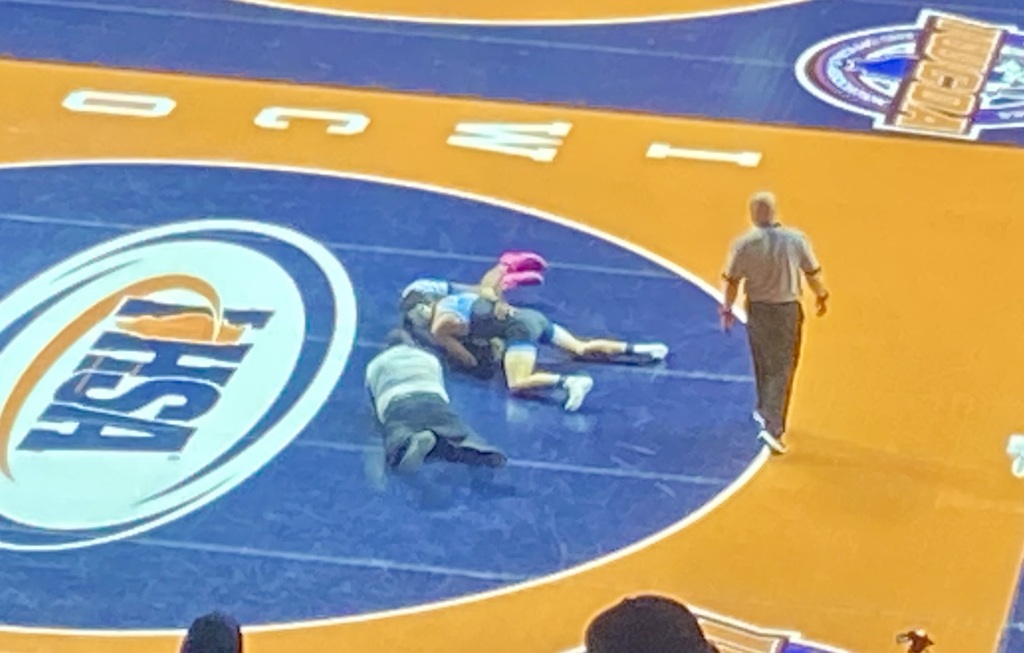 OM has pinned his opponent on the right side of the mat. One official is on the ground for the pin and the other is watching on top. 