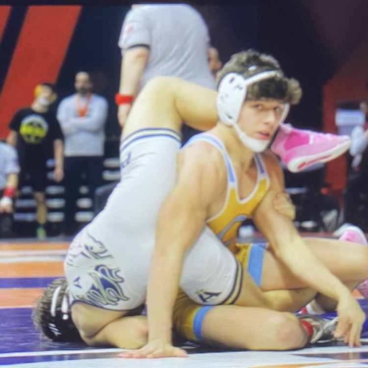 Owen is sitting up on the mat. His opponent is on his side with his head to the mat and his legs wrapped around Owen‘s waist and shoulder.