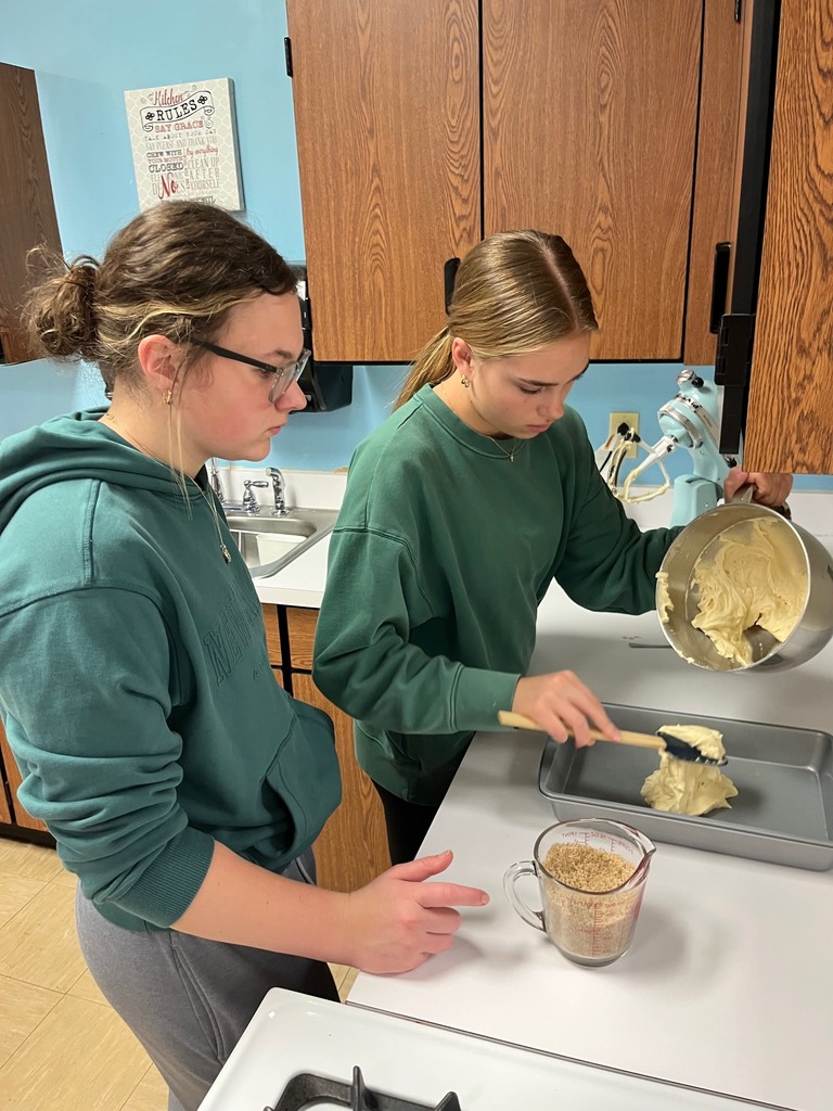 Two students are removing the batter from the bowl to the baking pan. There is about two cups of it in the middle of the pan.