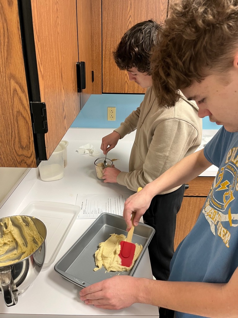 One student is flattening the batter with a spatula in the baking pan. Another student is mixing the toppings for the cake.