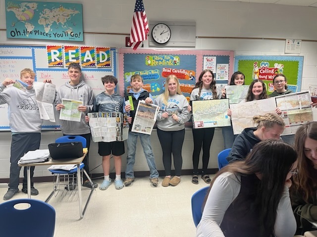 Nine students are standing in the back of the room holding up brochures that they have received from the parks. 