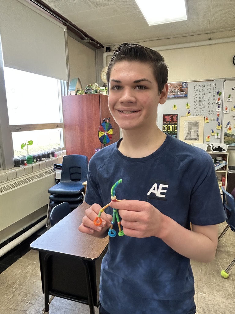A student is standing and holding his pipecleaner buddy.  It is made from pipecleaners and beads. 