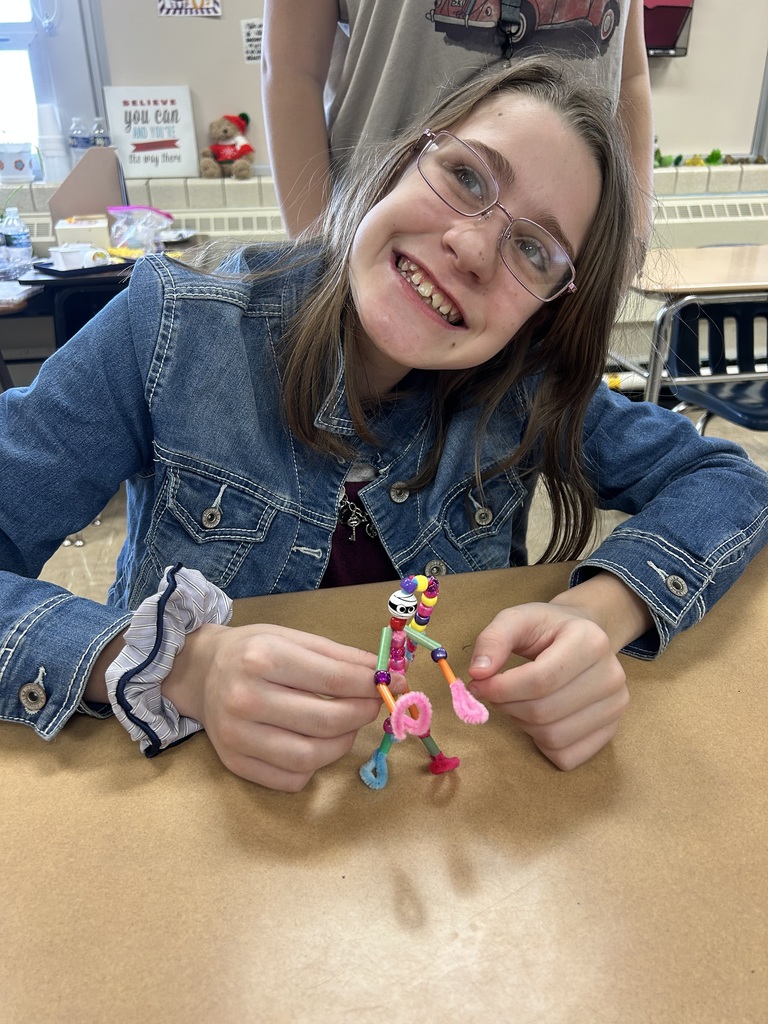 A student is sitting and holding her pipecleaner buddy.  It is made from pipecleaners and beads. 