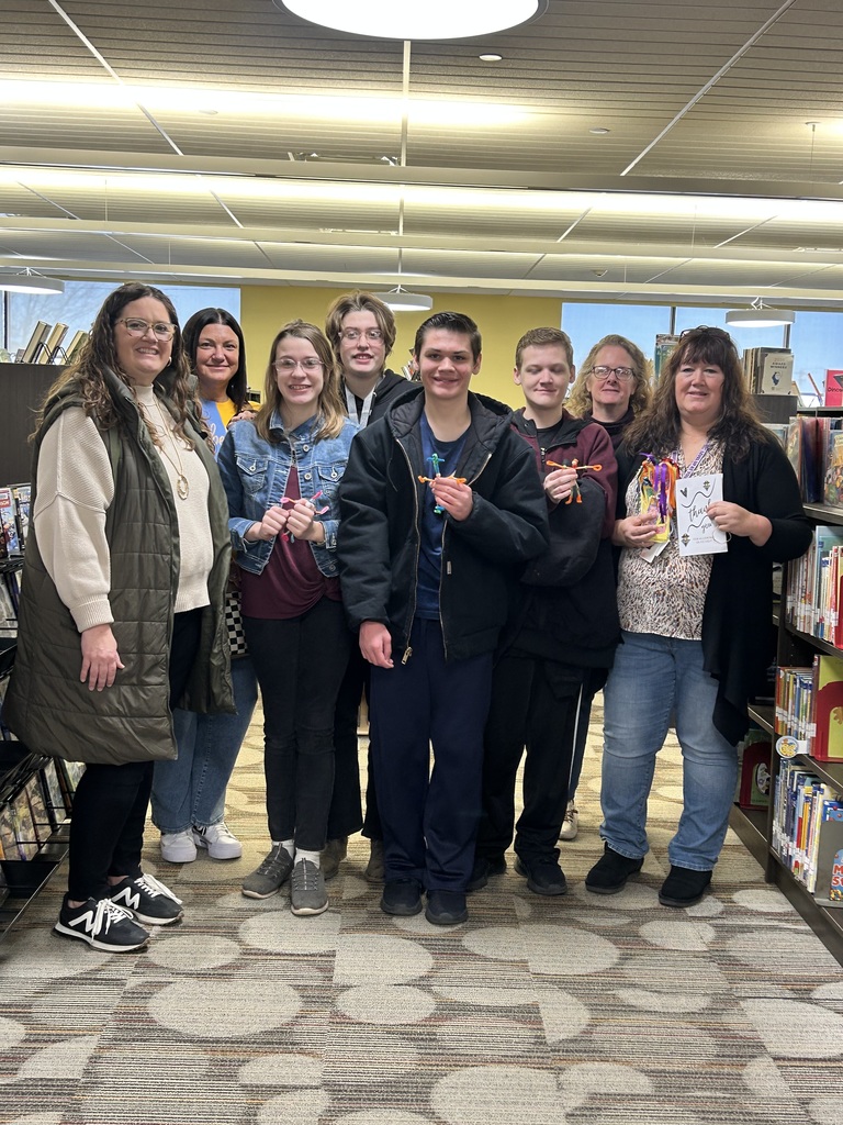 Students are standing between two bookshelves with the librarian and their teacher. 