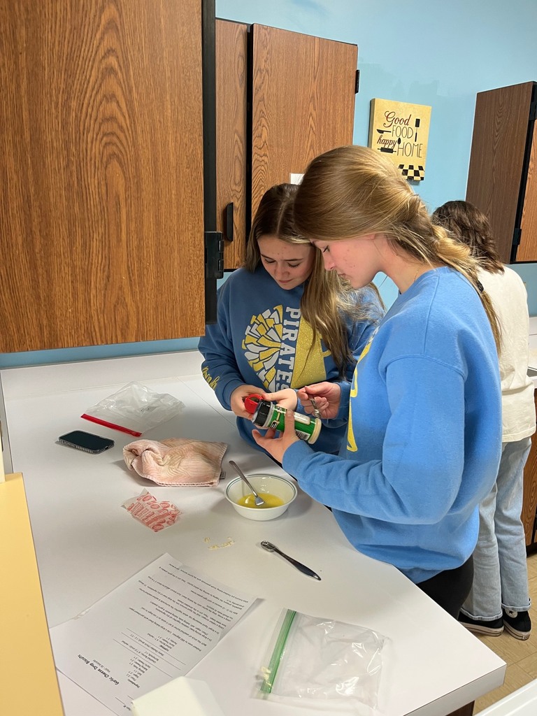 Two swtudents are looking at the backs of spice bottles. They have the recipe and a bowl of ingredients in front of them. 