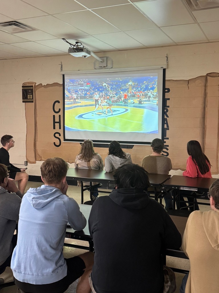 Students are in the cafeteria at the tables watching NB wrestle on the screen. 