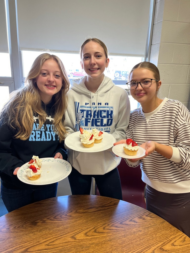 Three students are standing behind a table. They are each holding a plate of cakes iced with strawberries on top. 