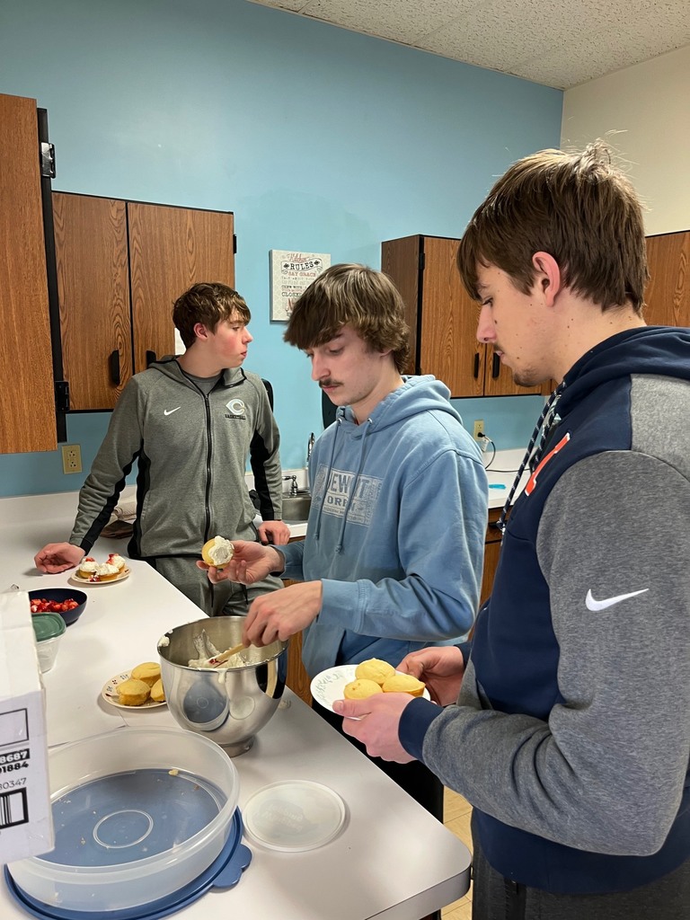 One student is getting icing out of a pan. Another is waiting in the background. A third is holding cakes to be iced. 