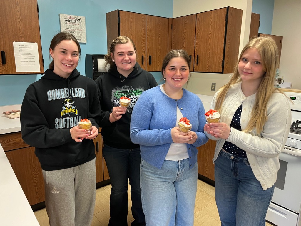 Four students are standing side by side each holding one iced cake with a strawberry on top. 