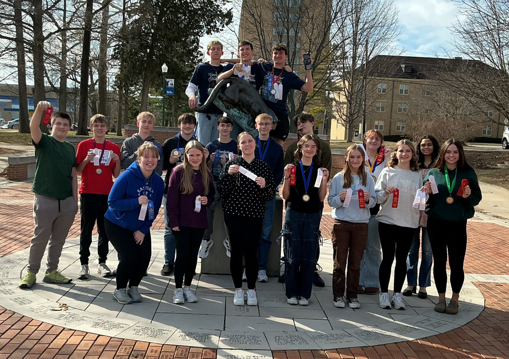 Pictured is the Math Team Standing in a group around and on a plant her statue. They are holidng ribbons and medals that they earned. 