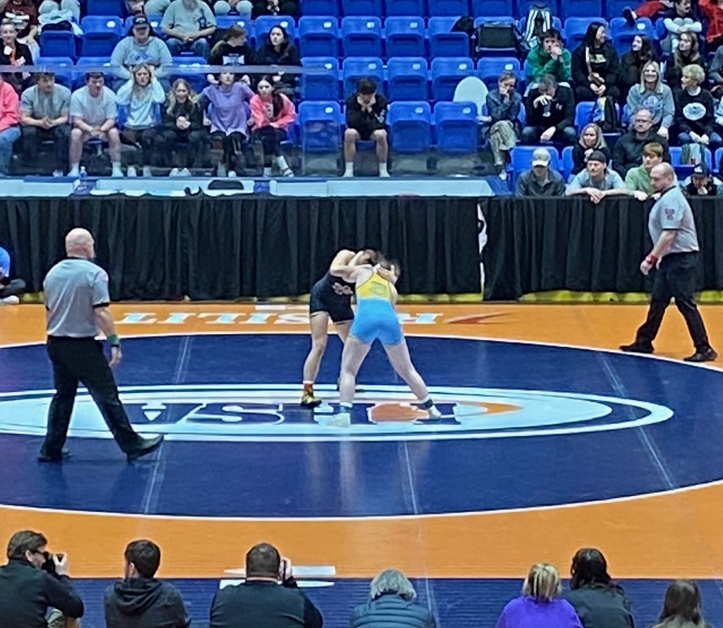 NB and opponent are standing with arms wrapped around the other's head and back. They are in the center of the mat. 