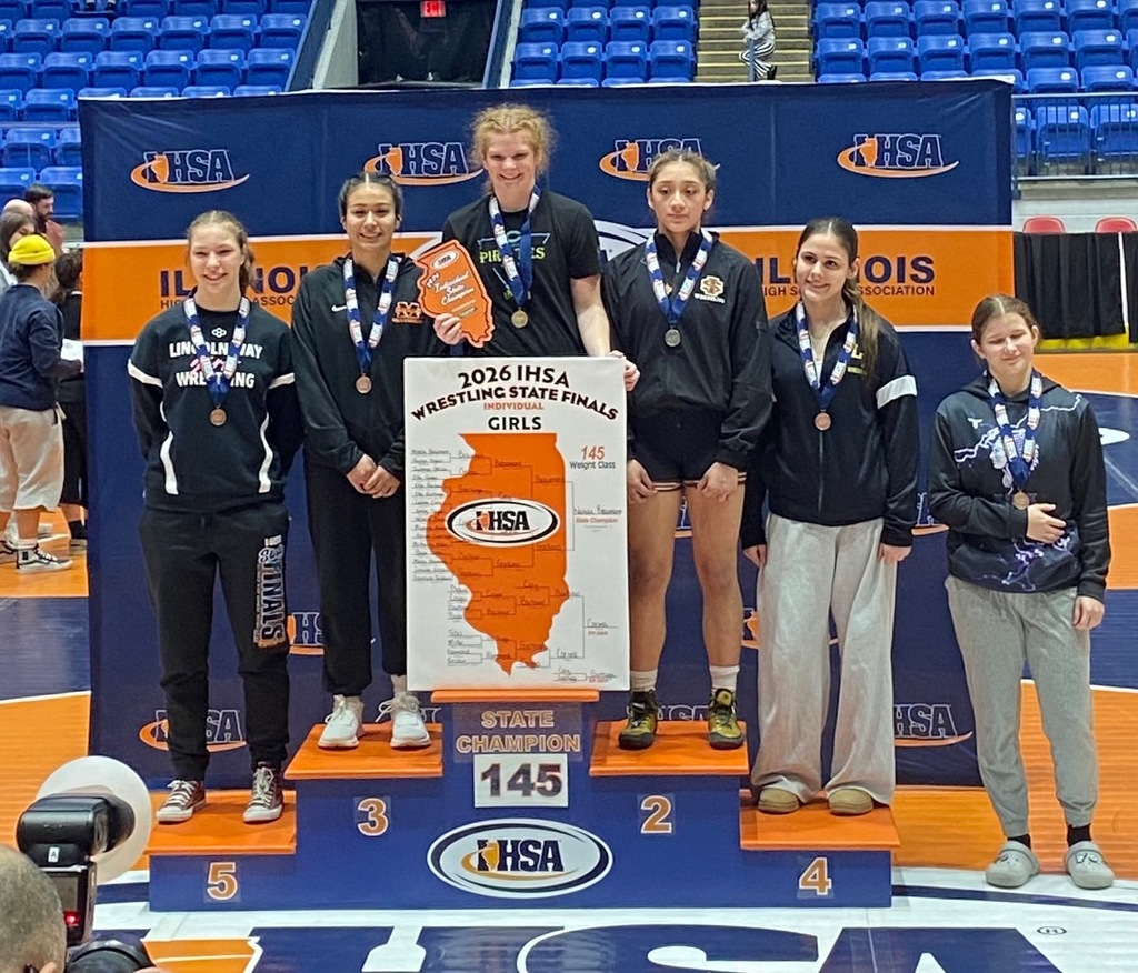 NB is standing on top of the podium. She is holding her individual champion trophy and bracket sign. She and the other other wrestlers are wearing their medals. 