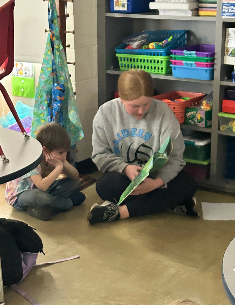 A middle school student is sitting next to an elementary school student on the  floor reading a book.
