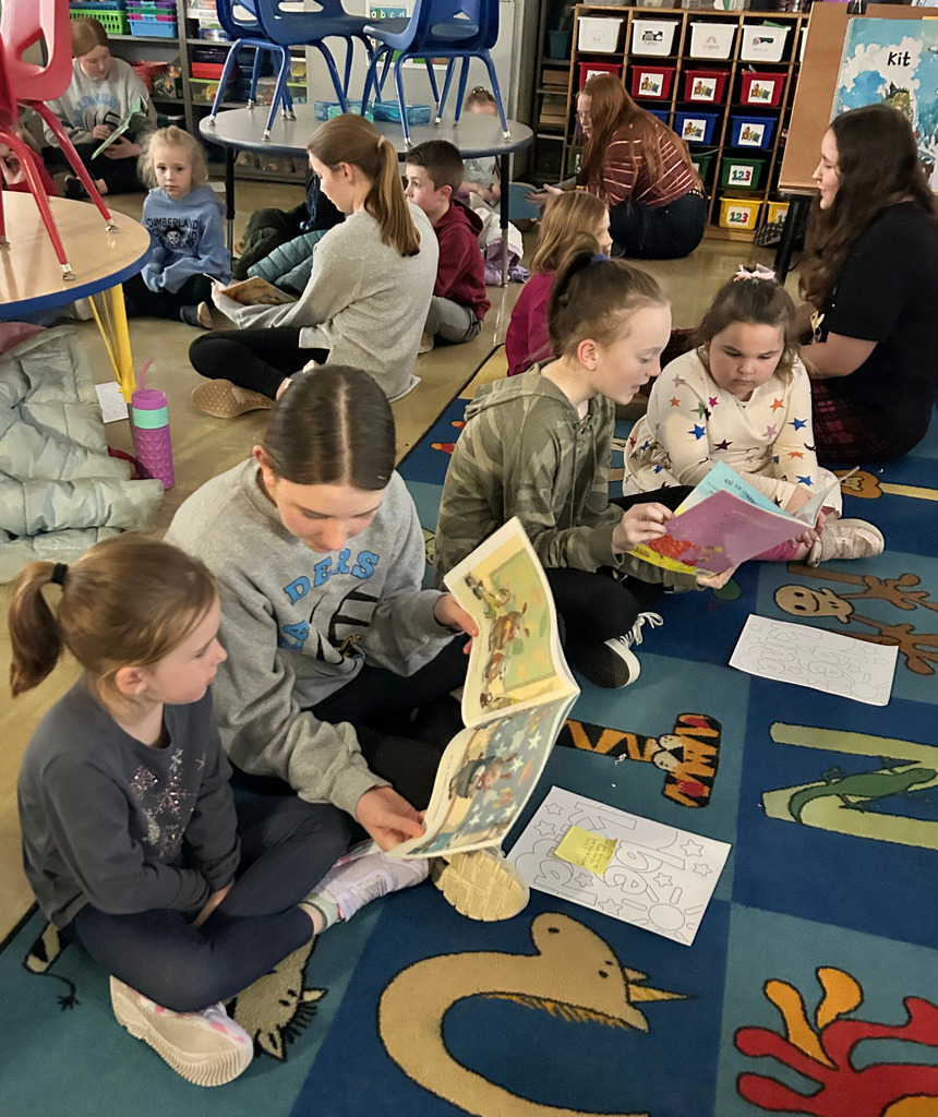 Middle school students are sitting next to an elementary school student on the  floor reading a book.