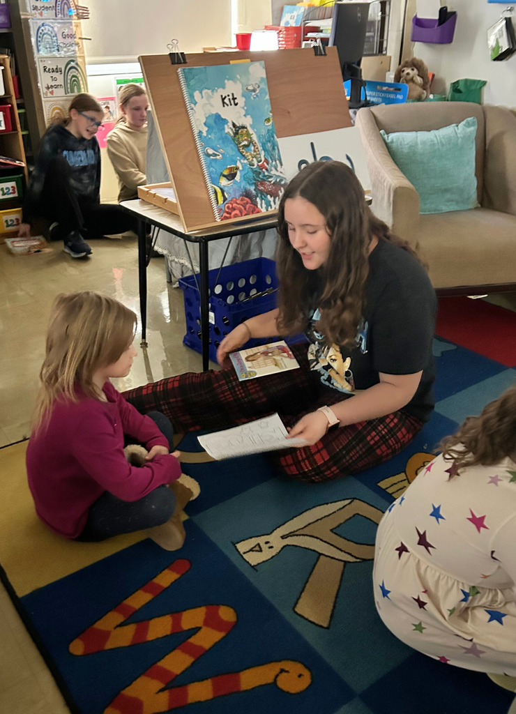 A middle school student is sitting next to an elementary school student on the  floor reading a book.