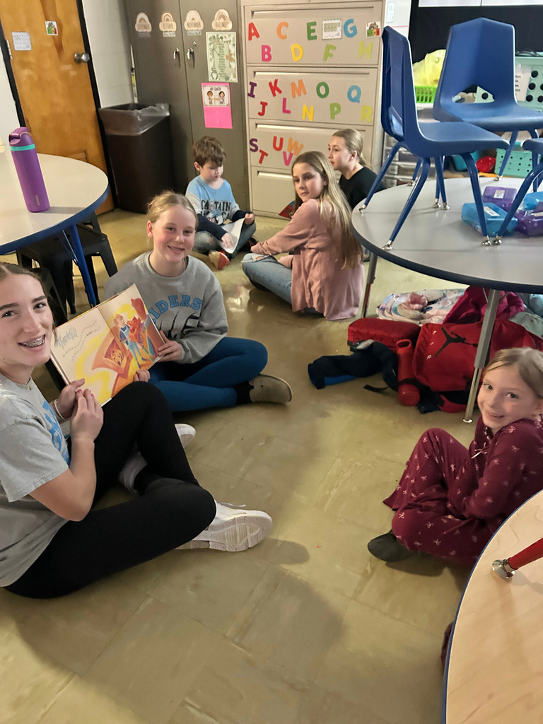 Middle school students are sitting next to an elementary school student on the  floor reading a book.