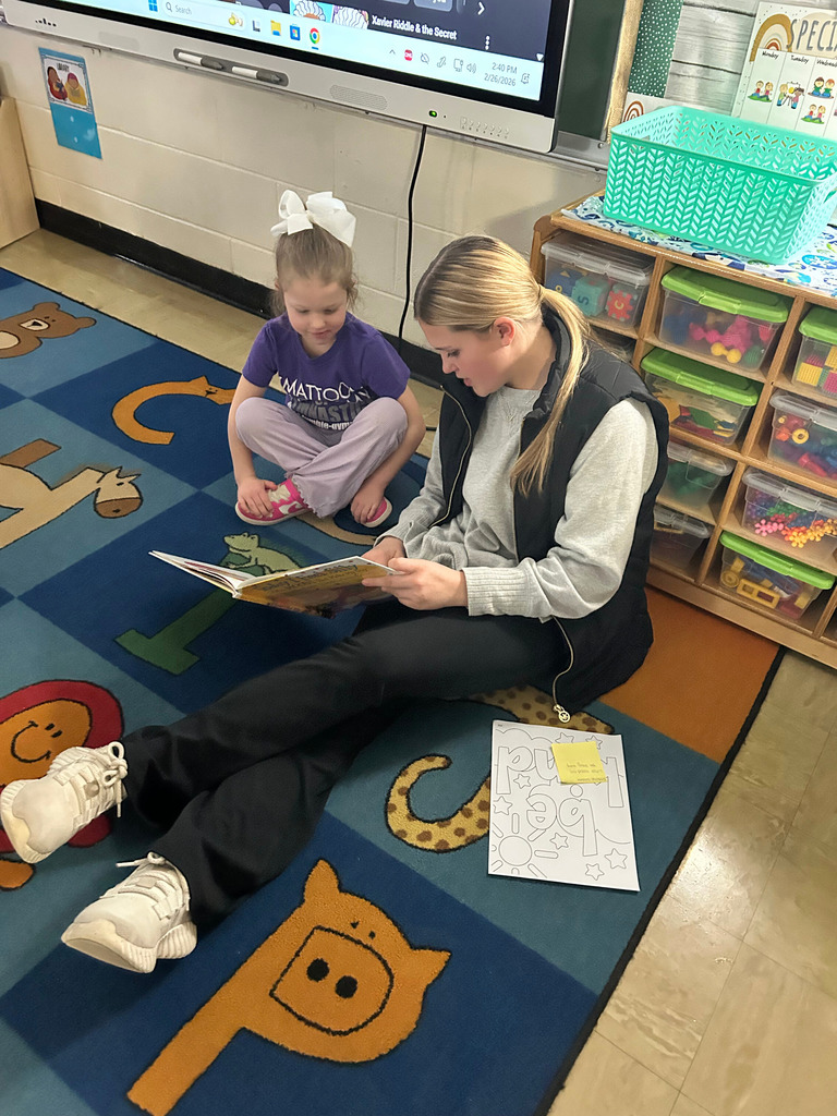 A middle school student is sitting next to an elementary school student on the  floor reading a book.