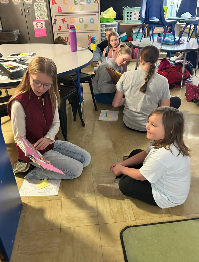Middle school students are sitting next to an elementary school student on the  floor reading a book.