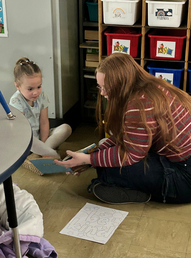 A middle school student is sitting next to an elementary school student on the  floor reading a book.