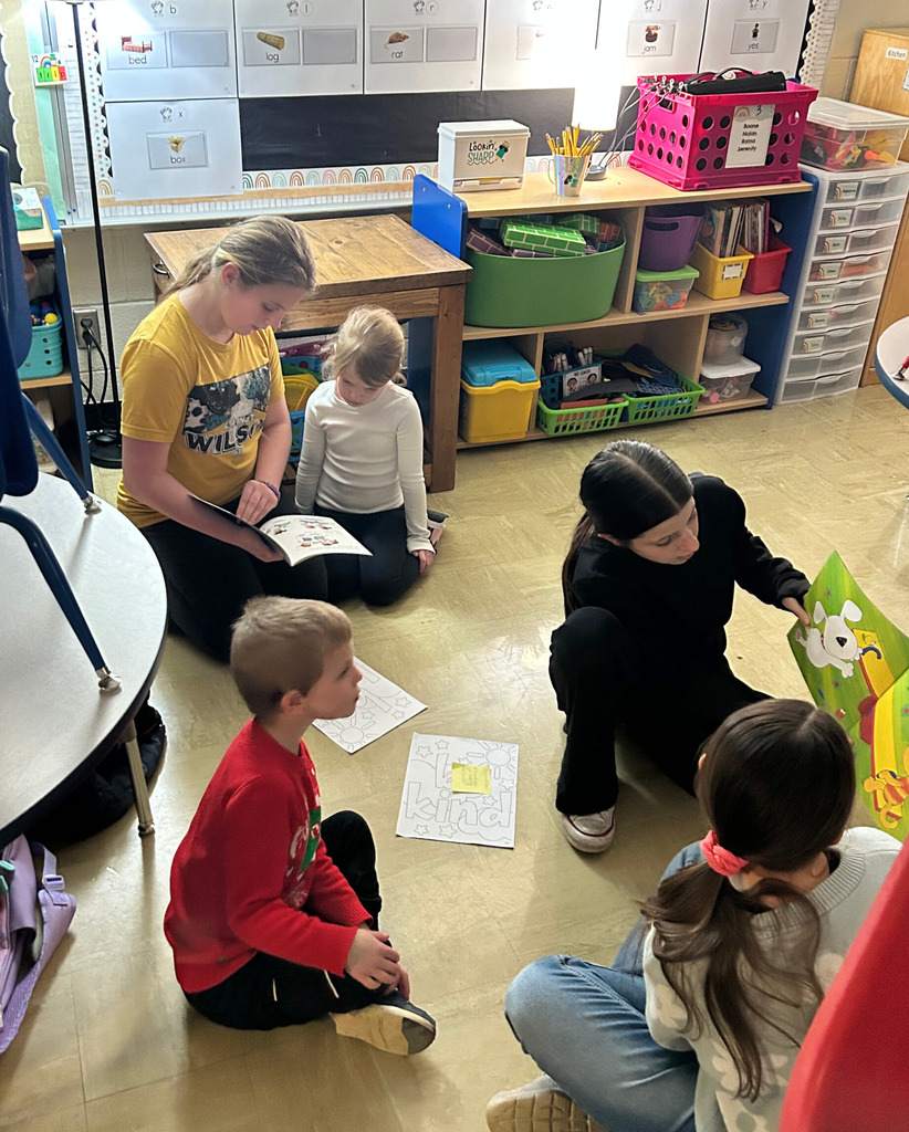 Middle school students are sitting next to an elementary school student on the  floor reading a book.