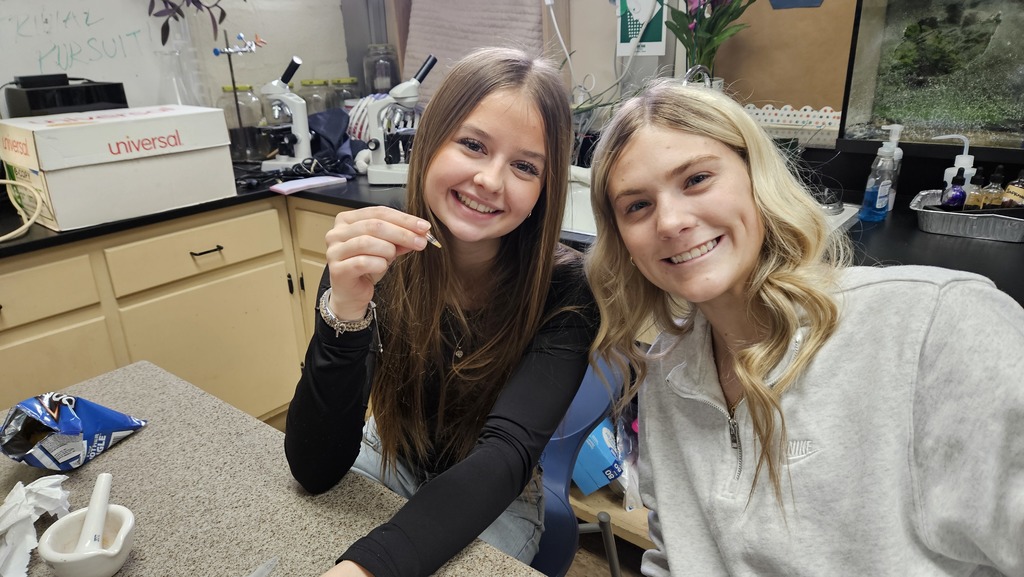 Two students are smiling at the camera. One is holding their testing material. 