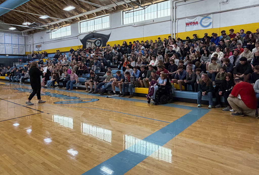 The student body is sitting in the bleachers. There is a speaker in the center of the basketball court reading a paper. 