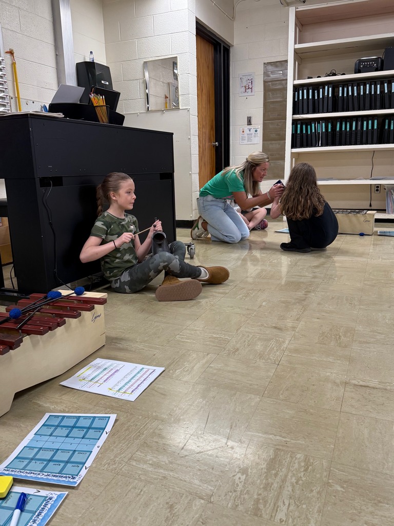 Two students are sitting on the ground and playing an instrument. A teacher is helping one student. 