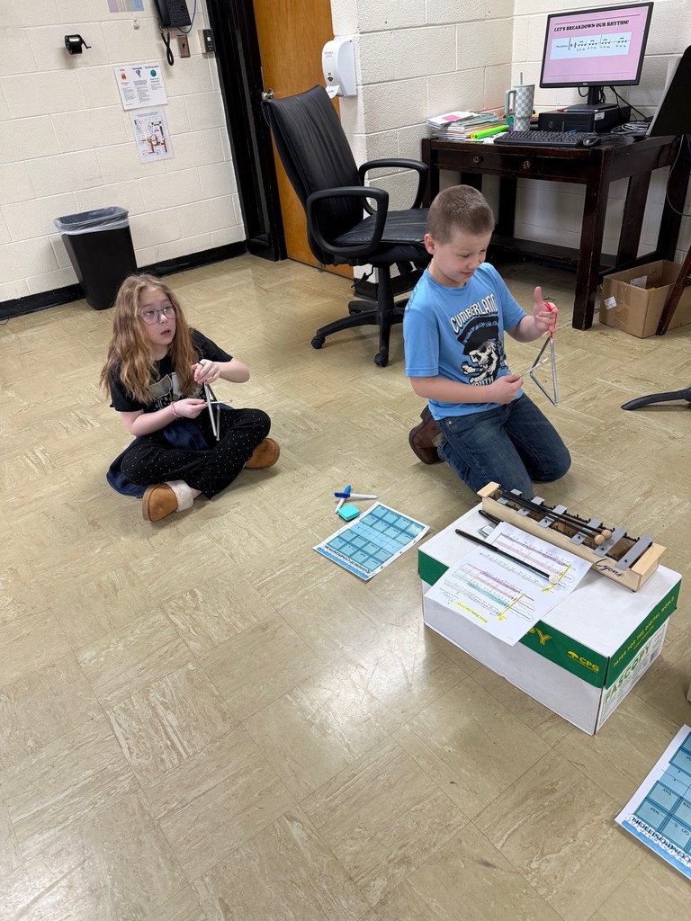 Two students are sitting on the ground and playing the triangle. 