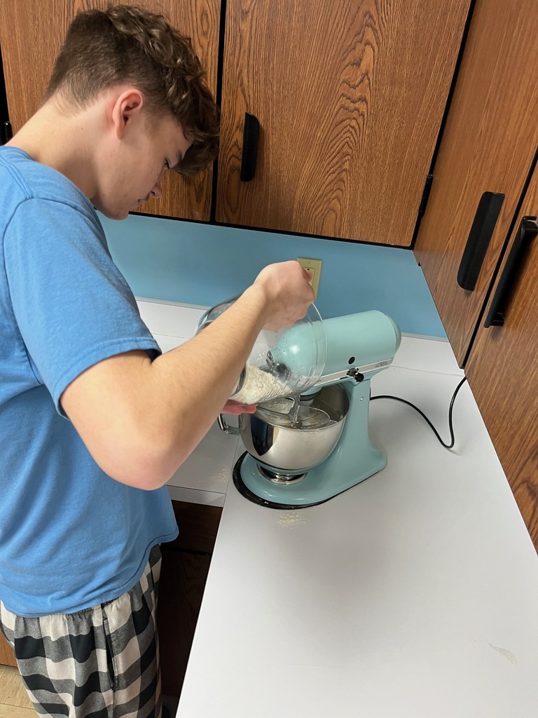 A student is adding ingredients to the mixing bowl and turning on the mixer. 