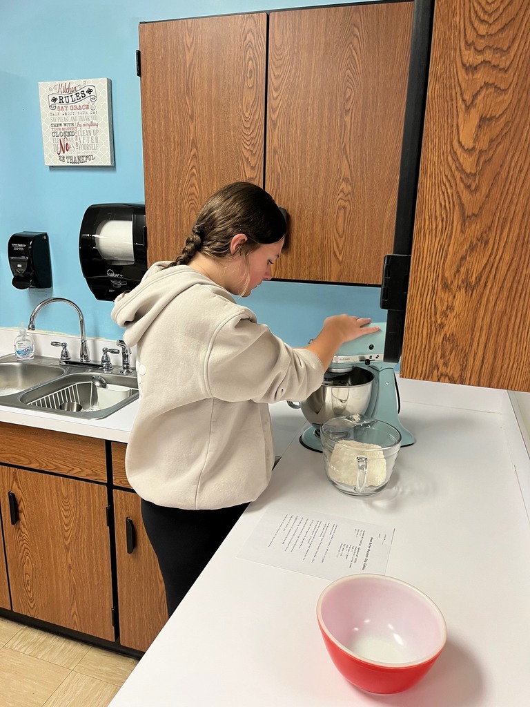 A student is adding ingredients to the mixing bowl and turning on the mixer. 